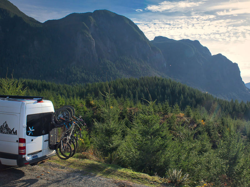 Tokul Racks Van with bicycles on a mountain road surrounded by trees and mountains.